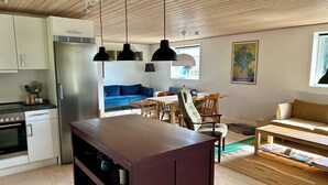 Una cocina moderna con horno empotrado, refrigerador y luces colgantes. Hay un comedor con mesa y sillas de madera, y un sofá azul al fondo.