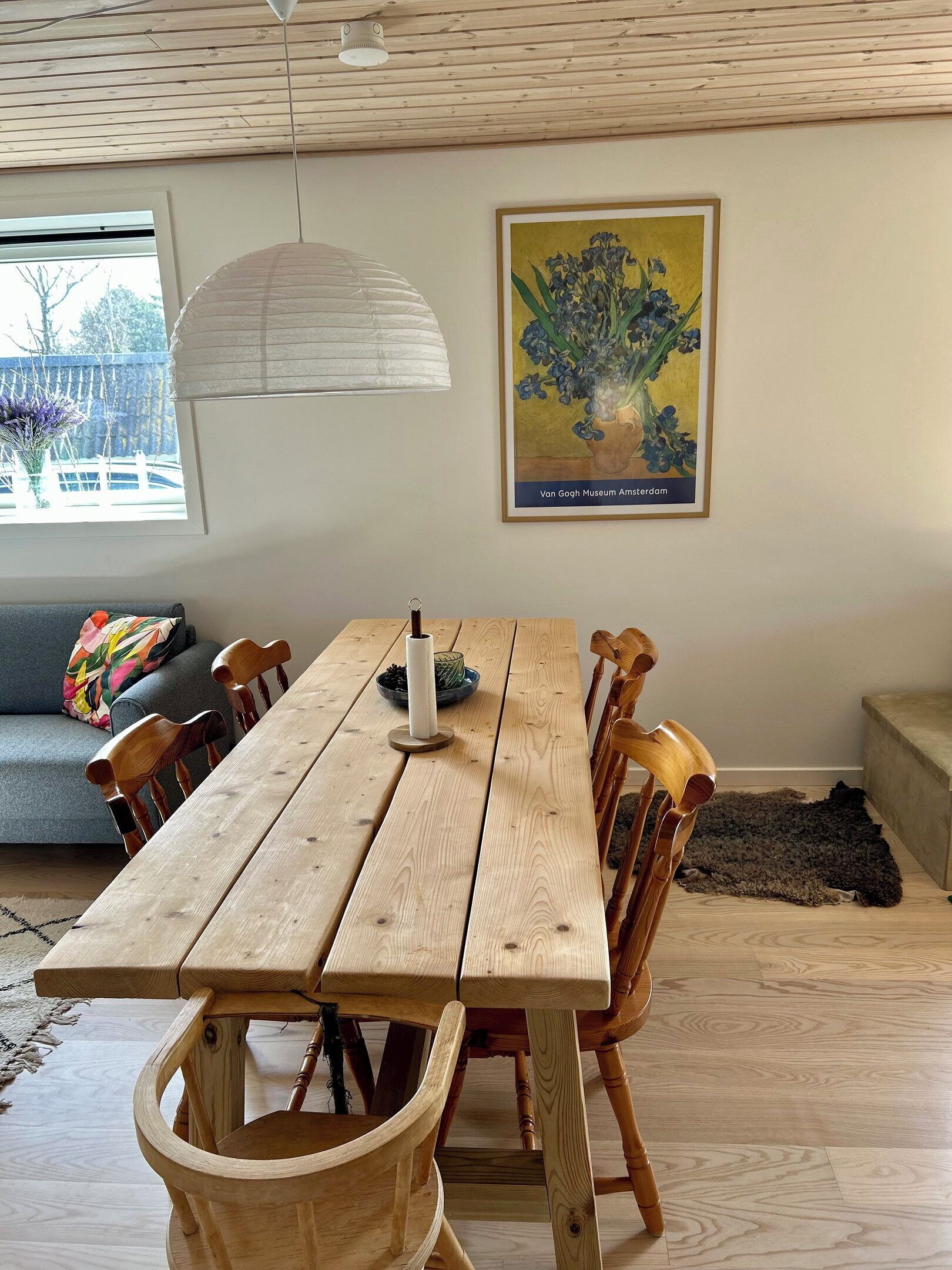 A wooden dining table with chairs, a hanging light fixture, a framed artwork, and a window with a view.