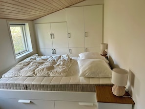 A bedroom with a bed, a bedside table with a lamp, a window, and a wardrobe.