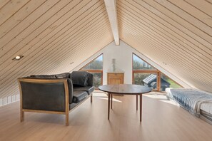 A high-ceilinged room with a sloped wooden roof, a sofa, a table, and a bed with a view of the outdoors. - Lovely cottage next to Dejbjerg Golf Club (Skjern)