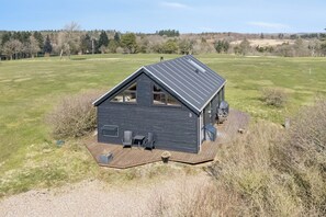 A modern house with a sloped roof, a deck with seating, and a view of a grassy field and trees in the background. - Lovely cottage next to Dejbjerg Golf Club (Skjern)