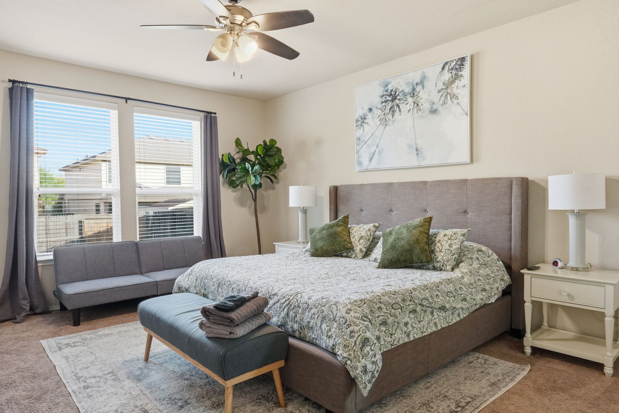 A bedroom with a bed, bedside tables, a bench, a ceiling fan, a window with curtains, and a wall-mounted artwork.