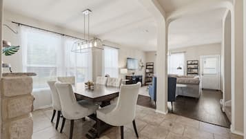A spacious dining area with a large table and chairs, a kitchen in the background, and a living area with a sofa and TV.