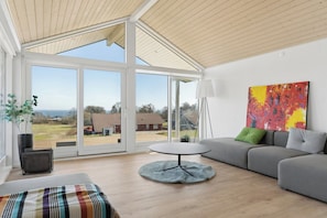 A modern living room with a large glass door leading to a balcony, a grey sofa, a round table, and a colorful abstract painting. - New 2026 holiday home with spa near beach (Augustenborg)