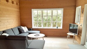 A wooden cabin interior with a gray sofa, a small round table, a TV on a stand, and a large window with a view of greenery. - Cozy log cabin near beach and forest (Strøby)