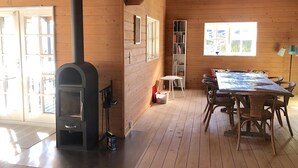 A wooden interior with a fireplace, a dining table with chairs, a bookshelf, and a window with a view. - Cozy log cabin near beach and forest (Strøby)