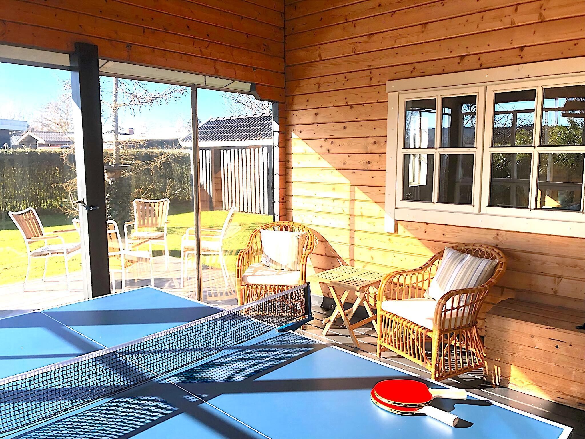 A wooden room with a ping pong table, wicker chairs, and a large window.