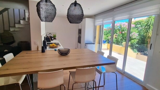 Comedor con mesa de madera, sillas blancas, dos lámparas colgantes y vista a un jardín a través de grandes ventanales.