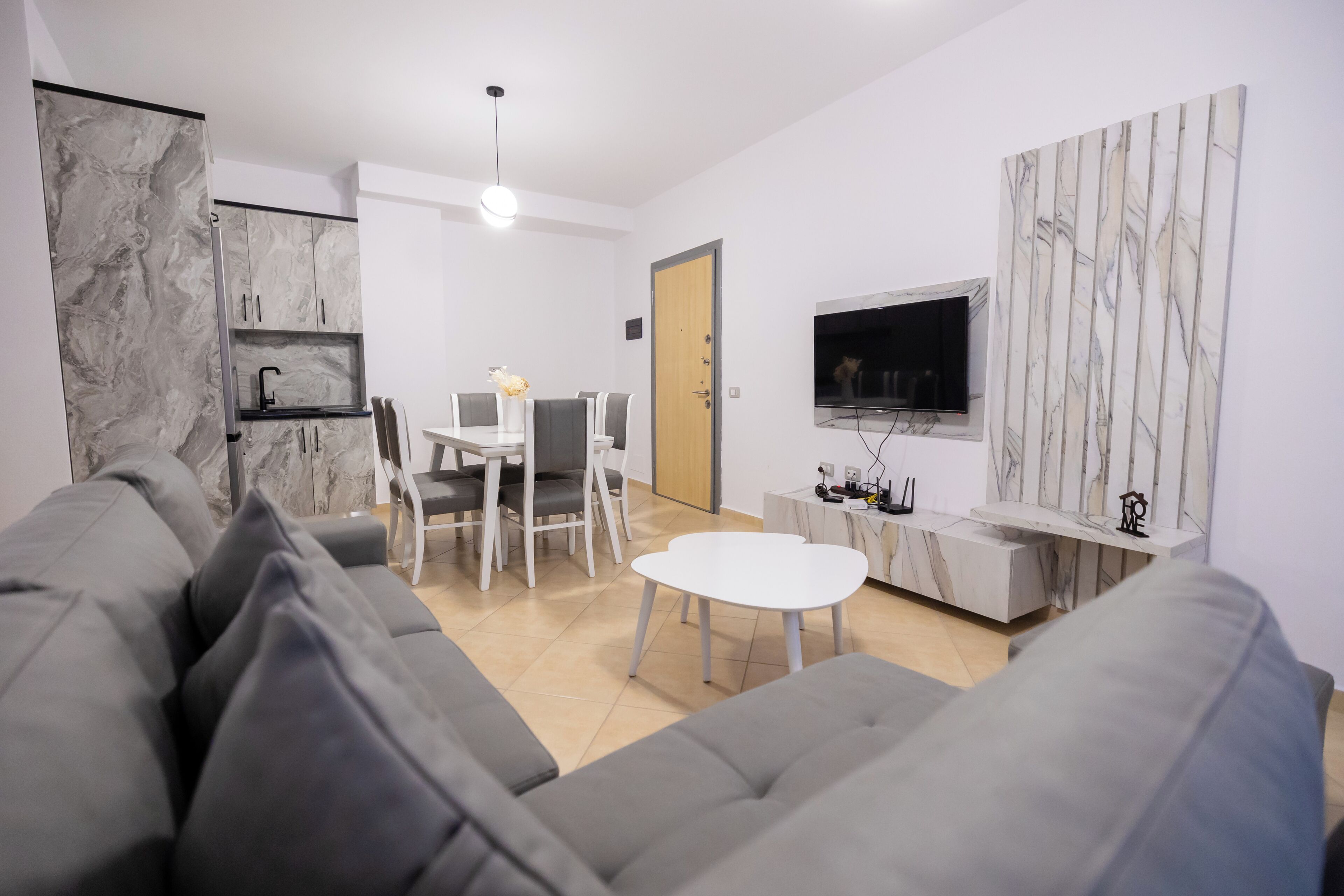 A modern living room with a grey sofa, a dining table with chairs, a white coffee table, a TV mounted on the wall, and a marble cabinet.
