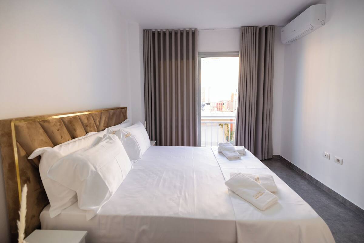 A hotel room with a bed, curtains, an air conditioning unit, and a view of the outside.