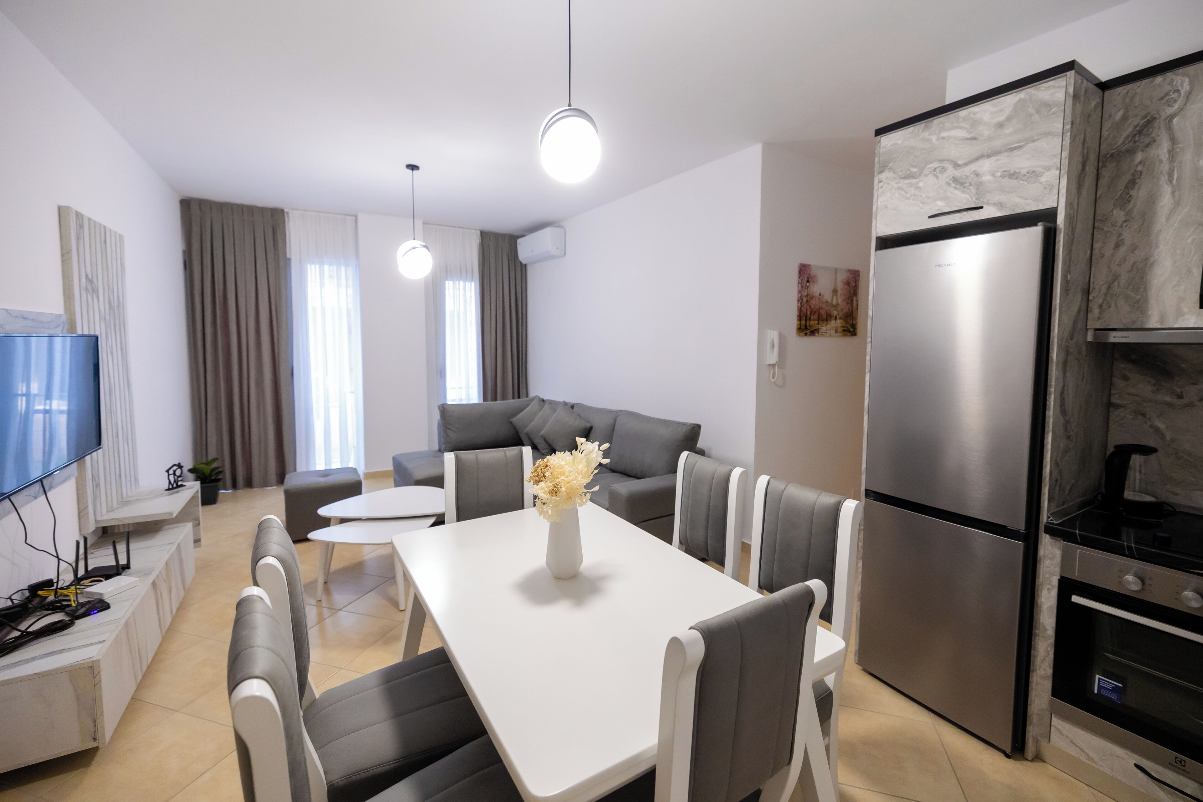 A modern kitchen with a stainless steel refrigerator, a dining table with chairs, a sofa, and a flat-screen TV.