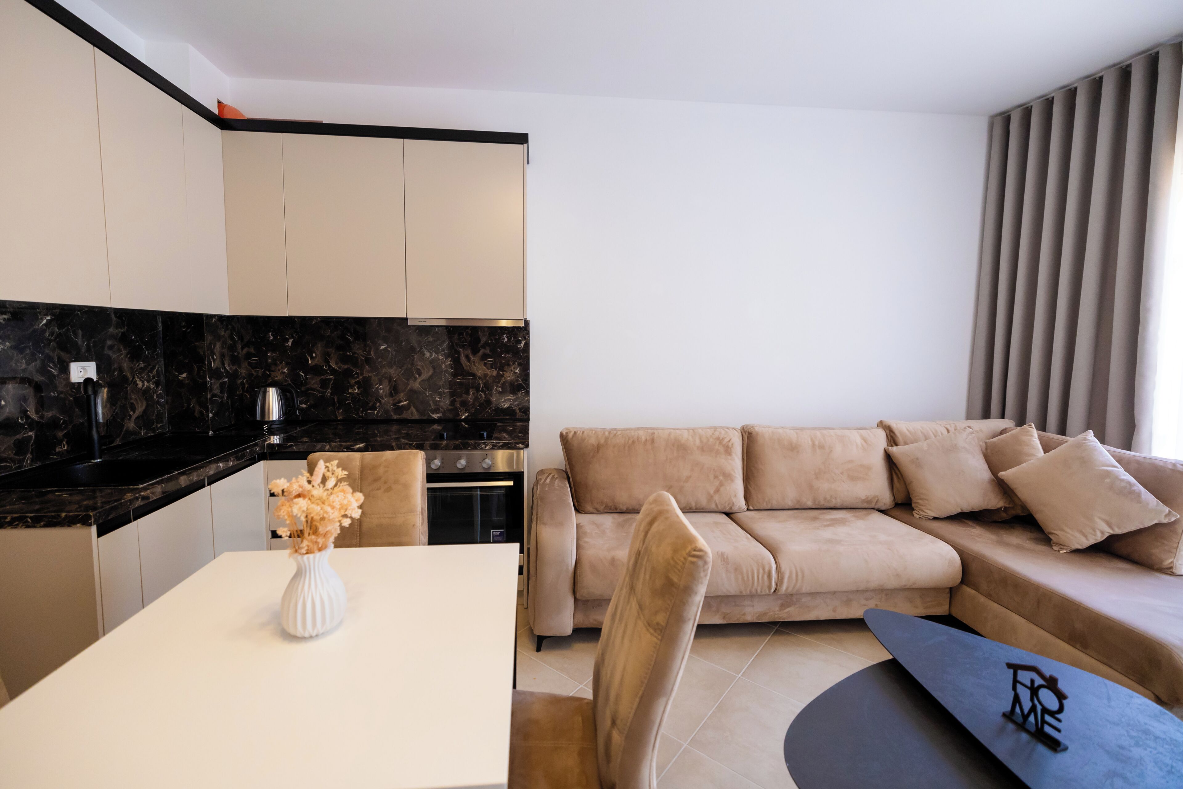 A modern living room with a beige sofa, a dining table with a vase, and a kitchen with black countertops.