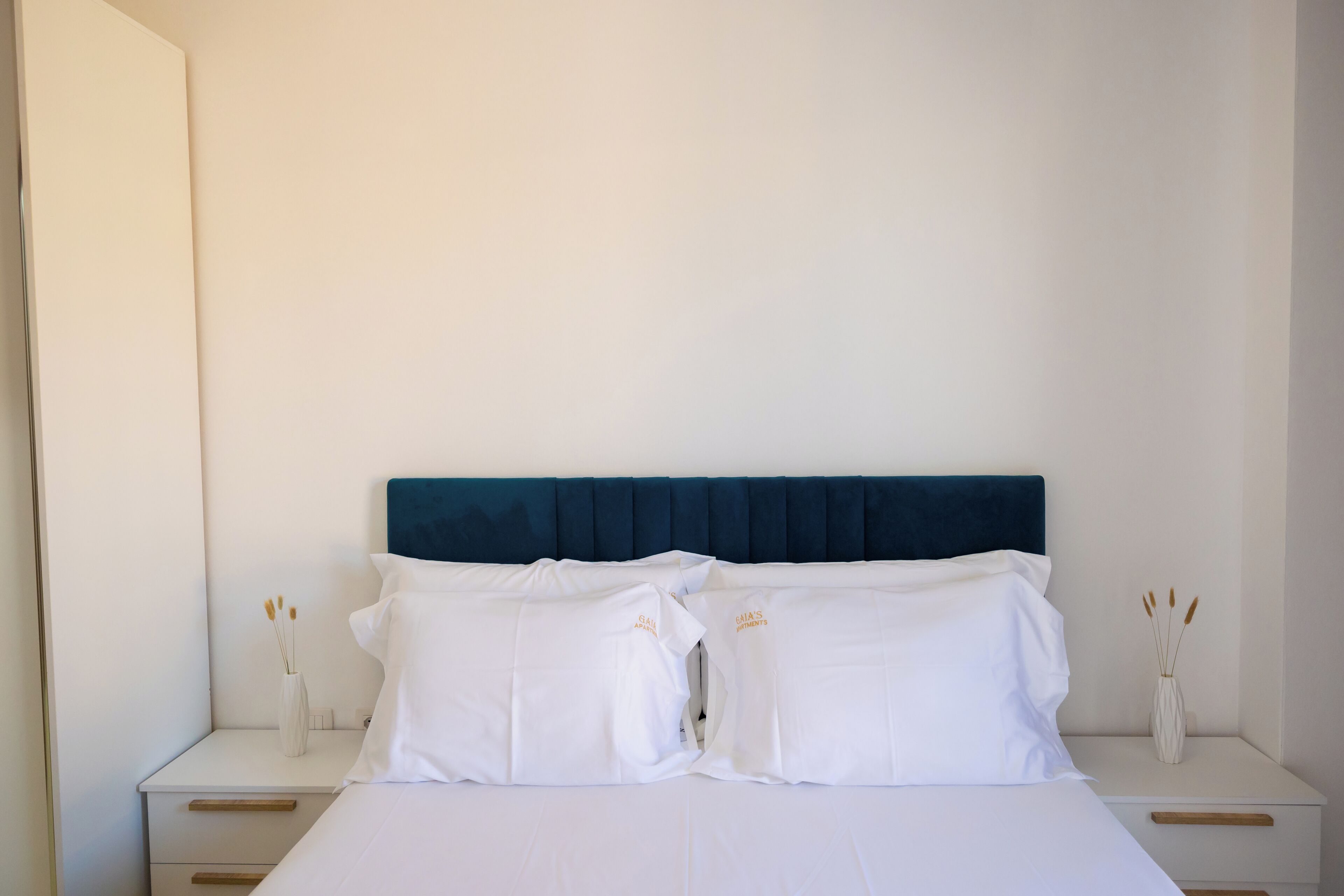 A neatly made bed with white linens and a dark blue headboard, flanked by white nightstands with vases of dried flowers.