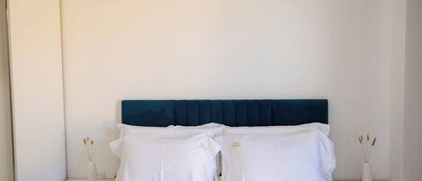A neatly made bed with white linens and a dark blue headboard, flanked by white nightstands with vases of dried flowers.