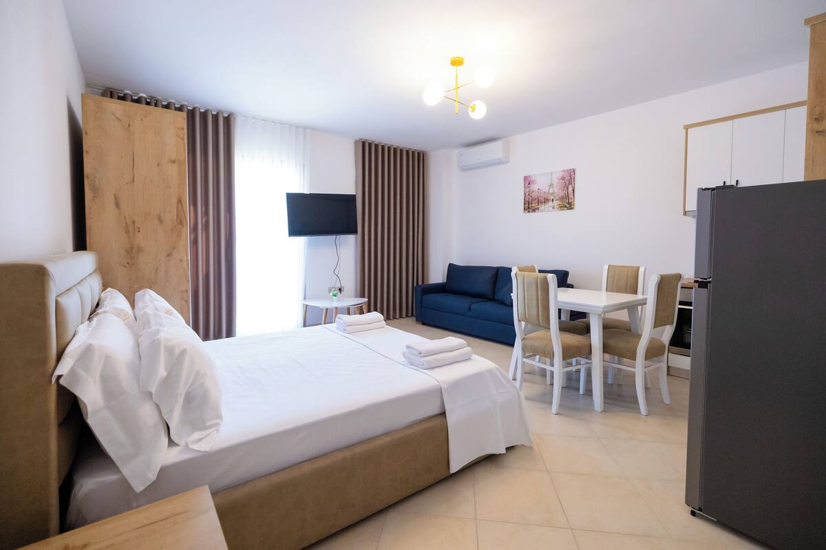 A hotel room with a bed, a dining table with chairs, a TV, and a refrigerator.