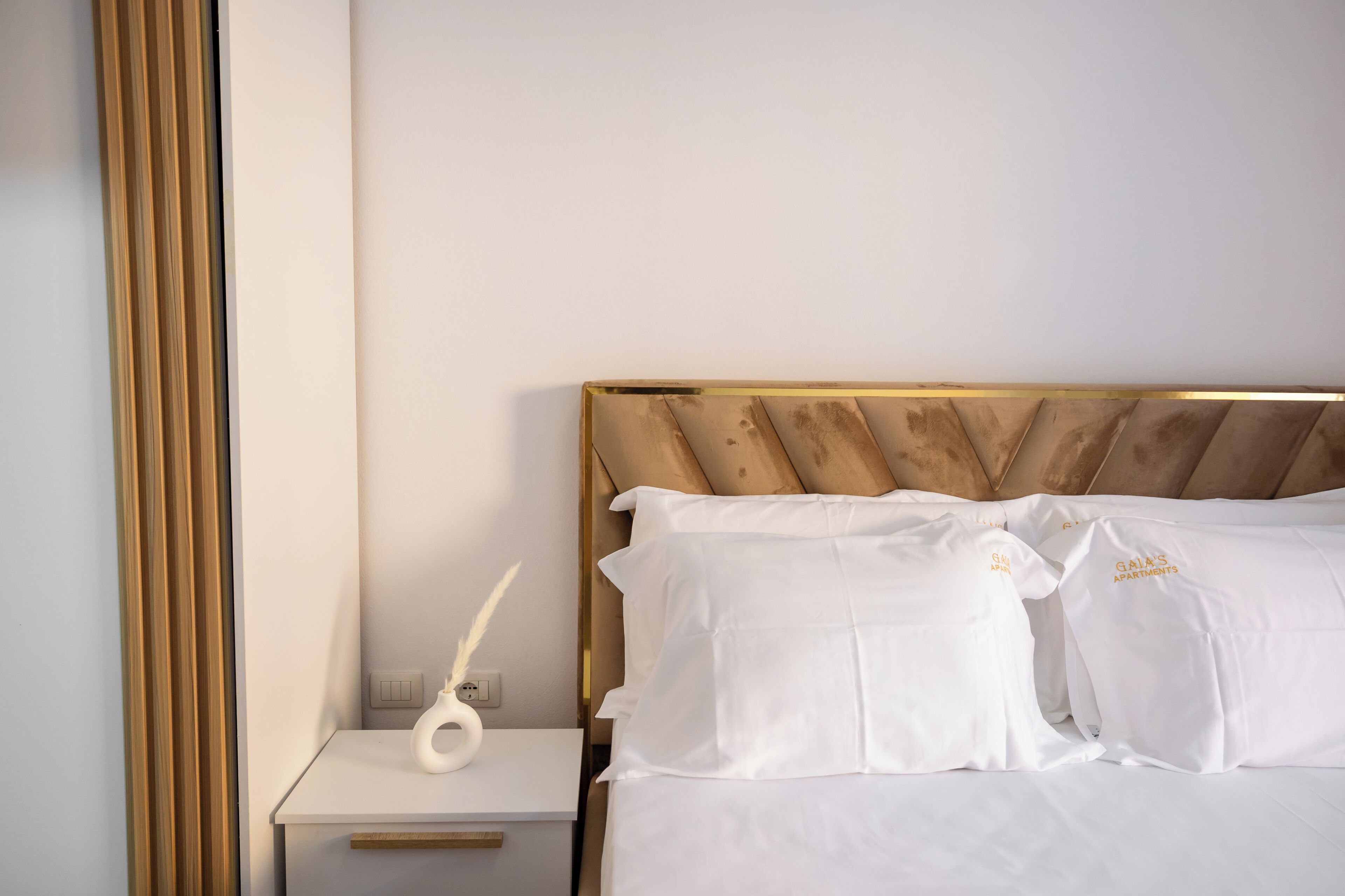 A neatly made bed with white linens and pillows, a bedside table with a white vase and a feather pen, and a wooden headboard.