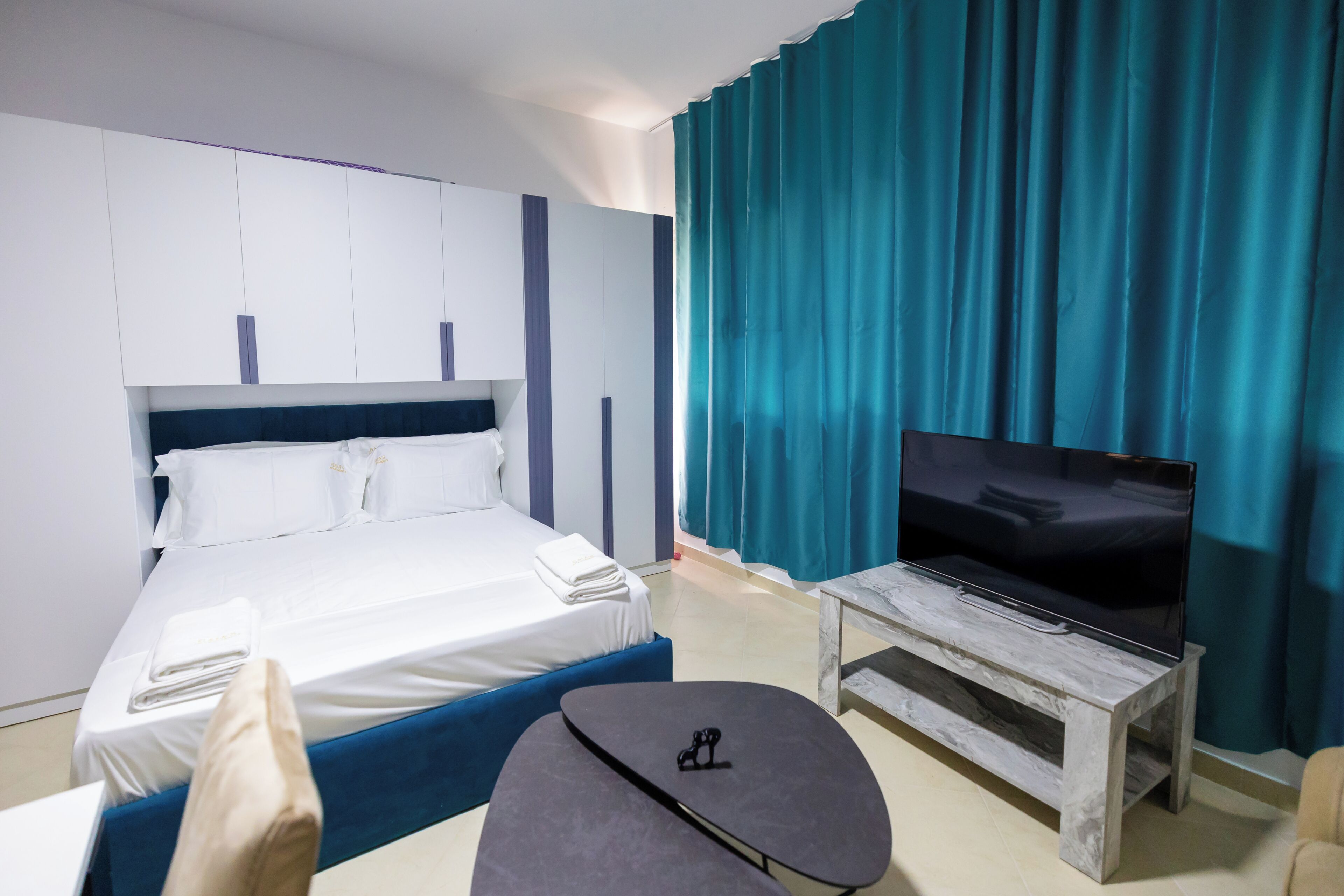 A hotel room with a bed, a flat-screen TV on a wooden stand, a small table, and a blue accent wall.