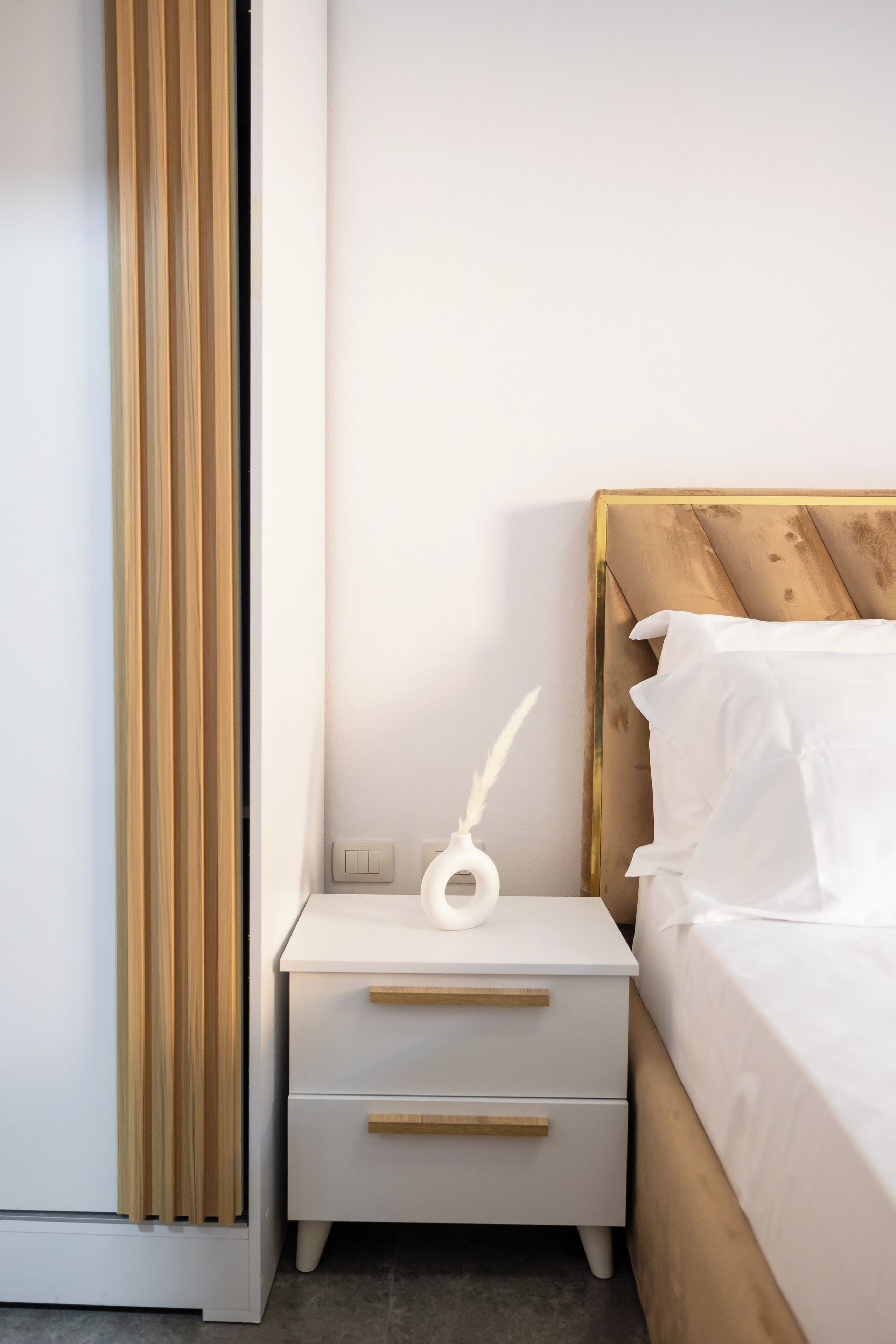 Une chambre à coucher avec une tête de lit en bois, du linge de lit blanc, une table de chevet avec un vase blanc et un tiroir.