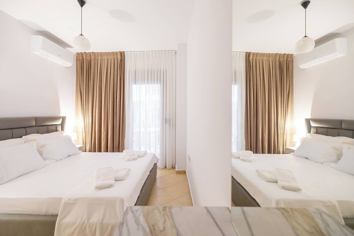 A hotel room with two beds, beige curtains, white bedding, and an air conditioning unit.