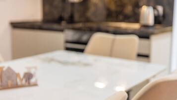 A modern dining area with a white table and beige chairs, a kitchen in the background with dark countertops, and a marble backsplash.