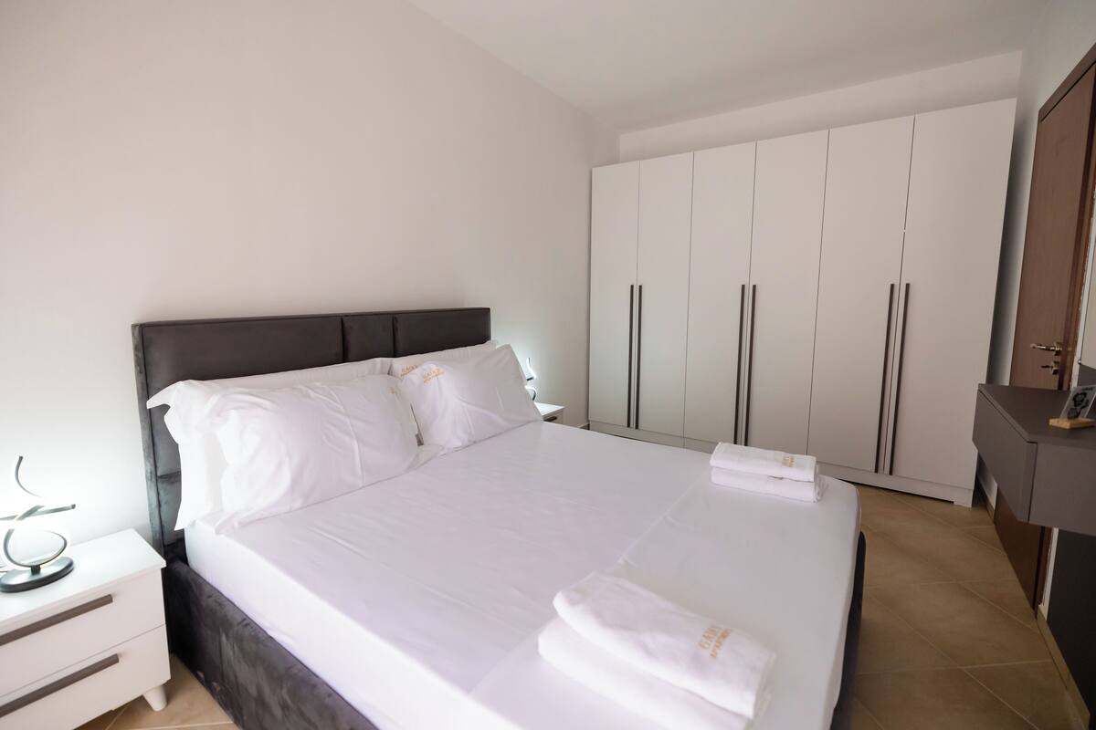 A neatly made bed with white linens, a dark headboard, and a white bedside table with a lamp.