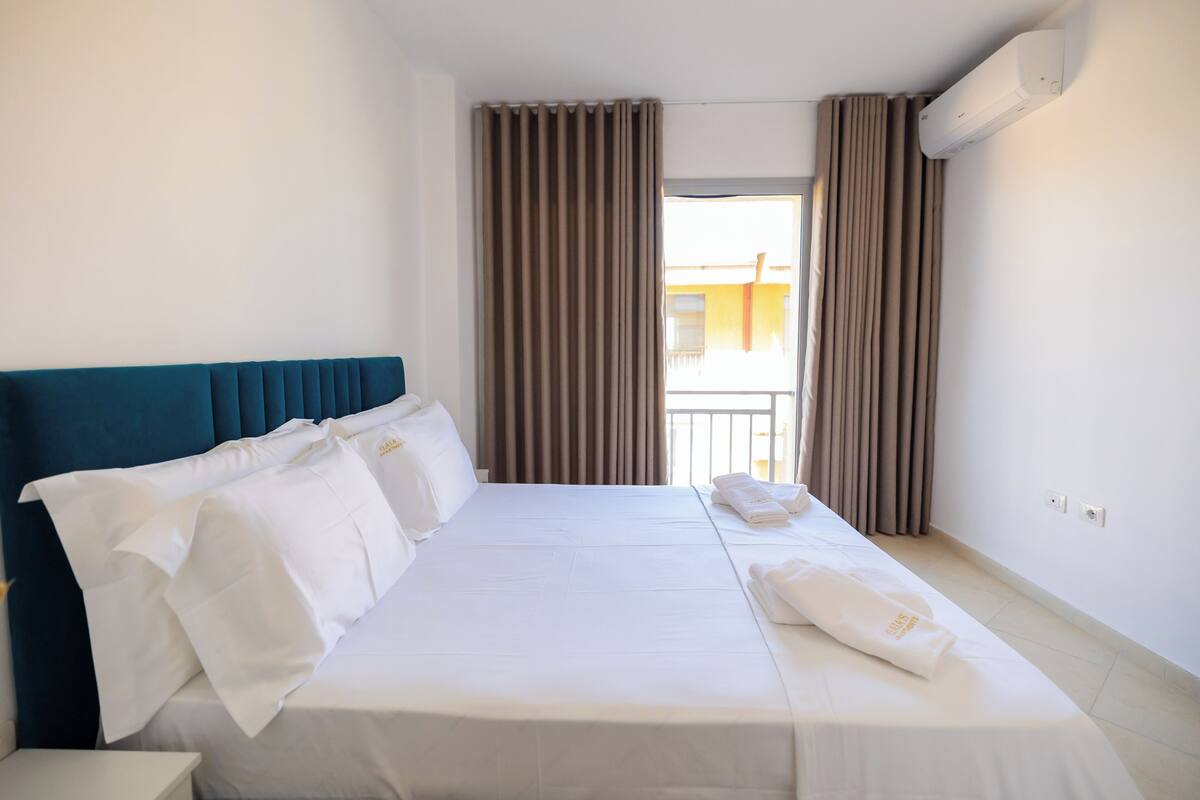 A hotel room with a bed, curtains, an air conditioning unit, and a view of a building outside.