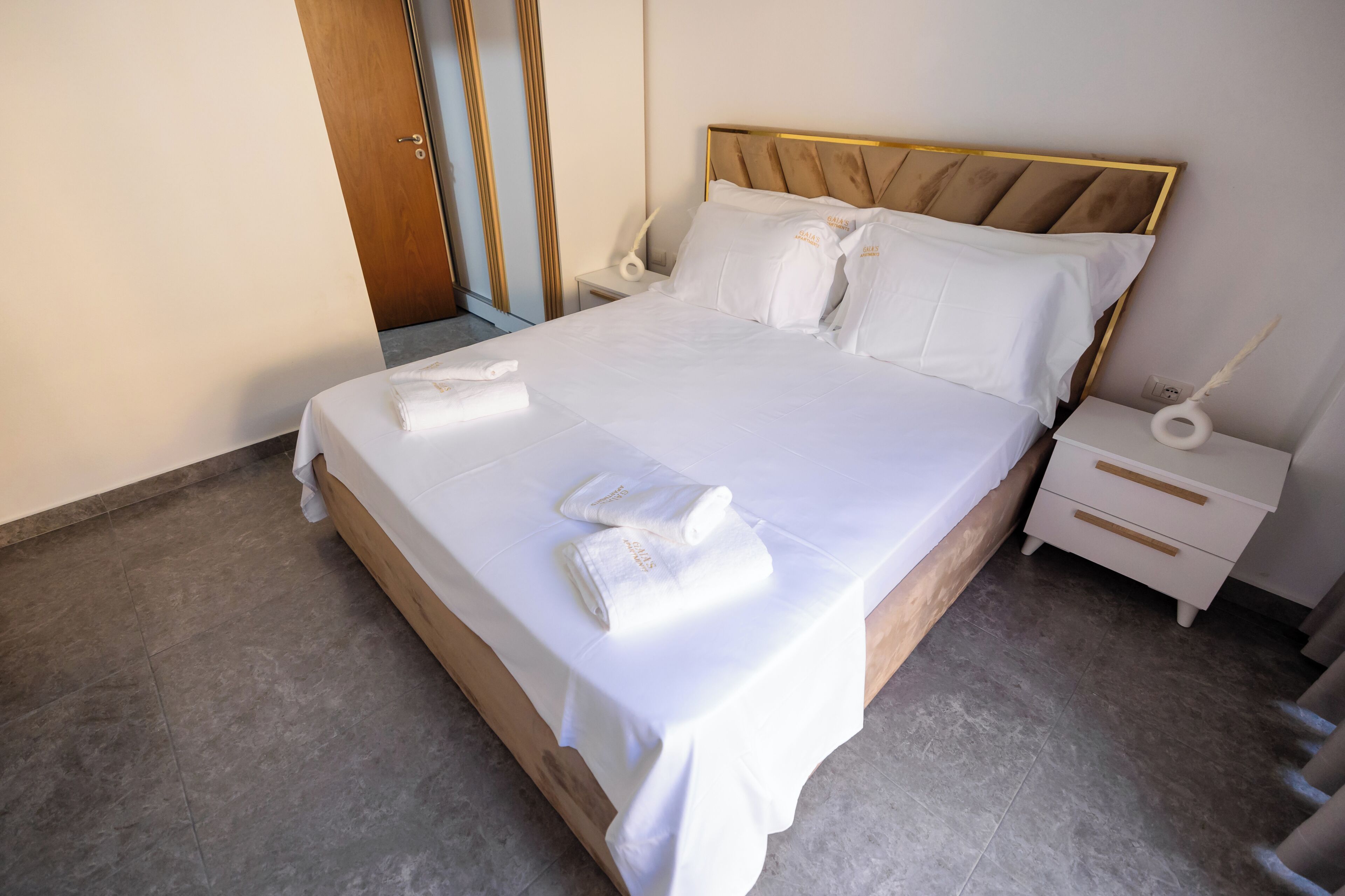 A neatly made bed with white linens and towels, a wooden headboard, a bedside table with a lamp, and a wall-mounted mirror.