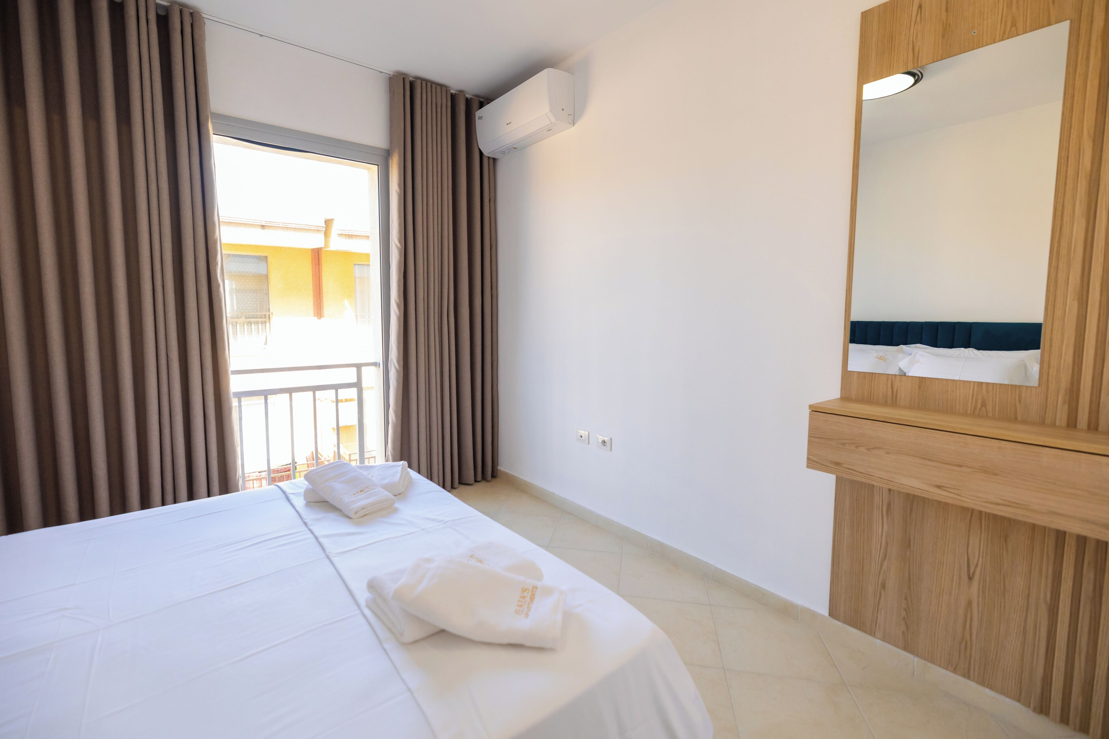 A hotel room with a bed, a mirror, a window with curtains, and an air conditioning unit.