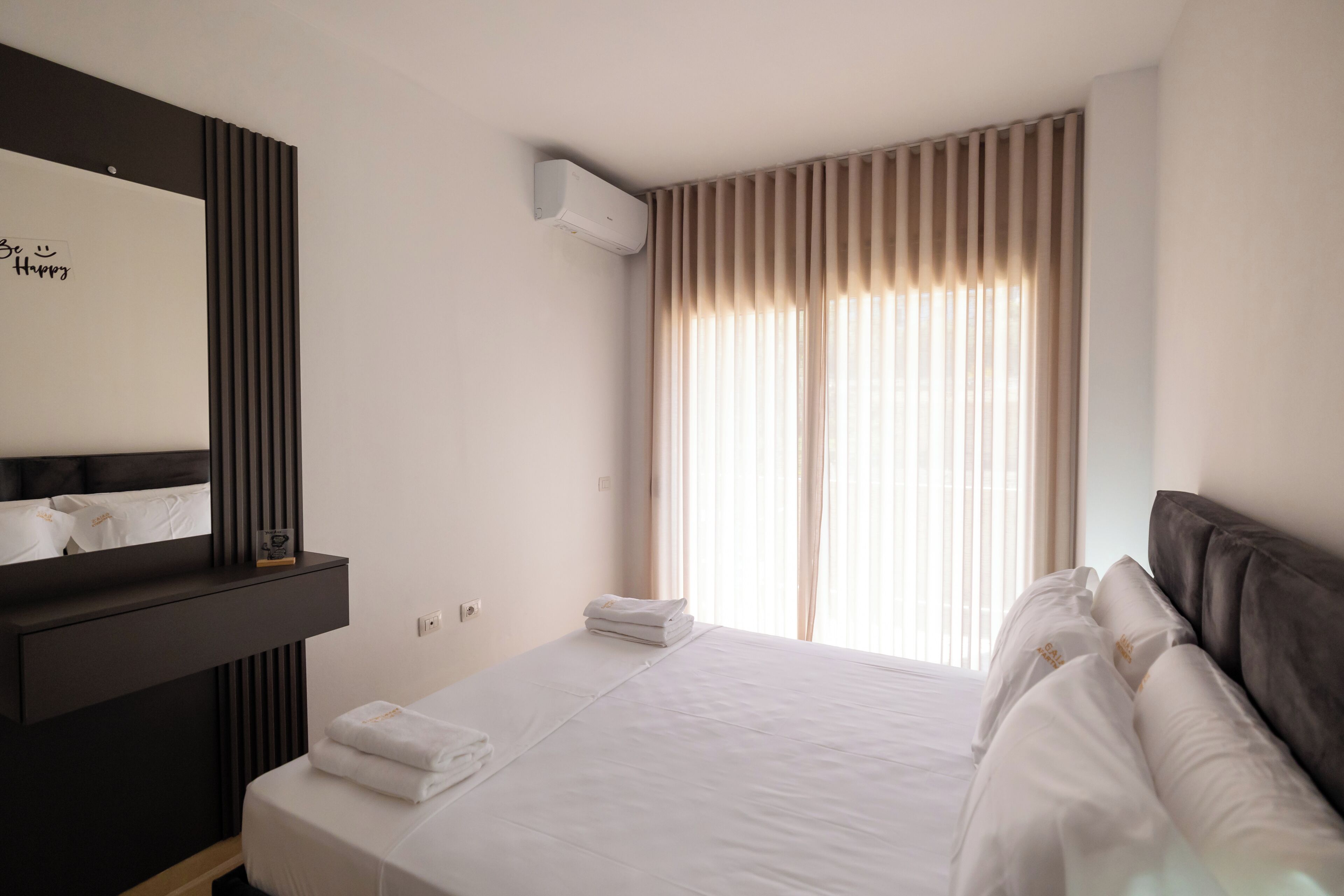A hotel room with a bed, a bedside table, a mirror, and a window with curtains.