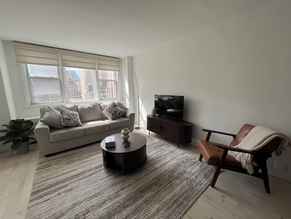 Un salon comprenant un canapé, une table basse, un téléviseur posé sur un meuble, un fauteuil et une grande fenêtre.