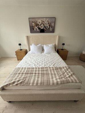 A bed with a striped blanket, two pillows, and a headboard. There are two bedside tables with lamps. A framed floral artwork is above the bed.