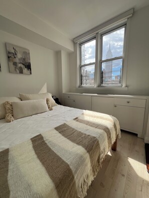 A bedroom with a bed, a window with a cityscape view, a radiator, and a painting on the wall.