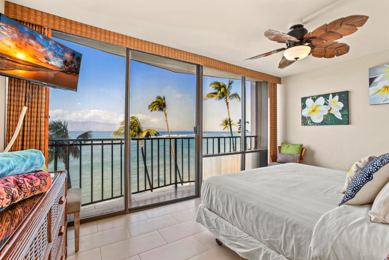 A bedroom with a large bed, a ceiling fan, a TV, and a balcony with a view of the ocean and palm trees.