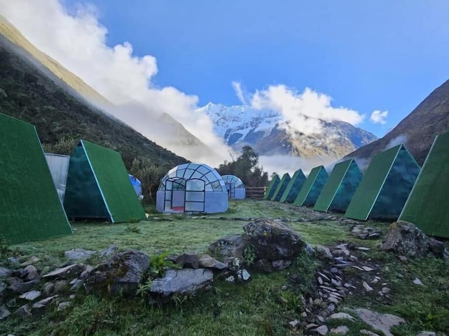 Várias tendas verdes numa área montanhosa, com céu claro e uma montanha coberta de neve ao fundo.