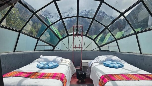 A room with two beds, a central table, and a view of mountains through a large window.