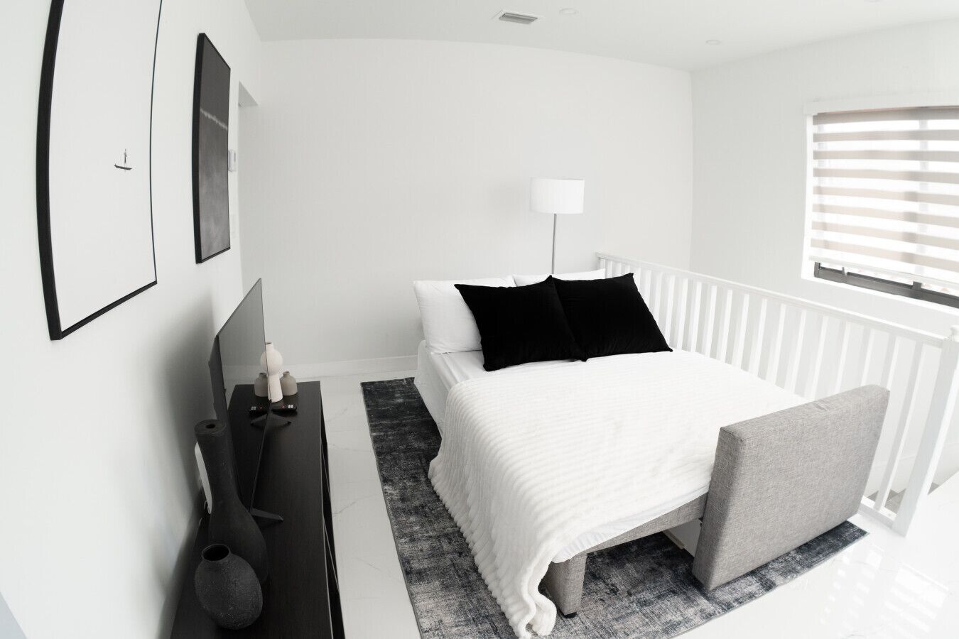A modern bedroom with a bed, bedside table, lamp, and a window with blinds.