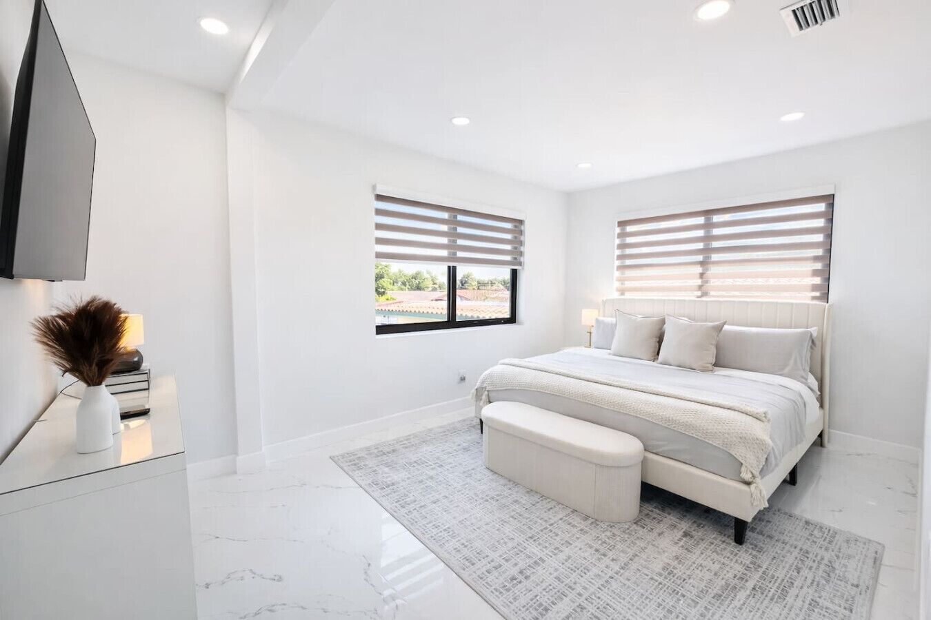A modern bedroom with a large bed, a bedside table, a TV, and two windows with blinds.