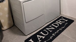 Une buanderie équipée d’un lave-linge et d’un sèche-linge, d’une petite étagère avec des articles de toilette et d’un tapis sur le sol, sur lequel est écrit le mot « Buanderie ».