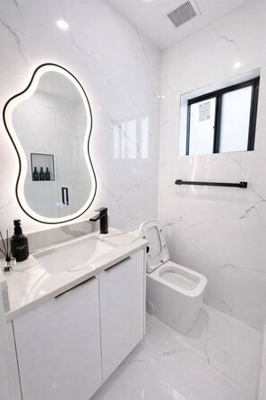 Ein modernes Badezimmer mit weißer Marmorwand, einem großen Spiegel mit schwarzem Rahmen, einer Toilette und einem Waschbecken mit schwarzem Seifenspender.