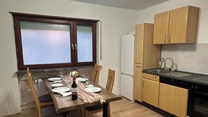 Een kleine keuken met houten kastjes, een eettafel voor vier personen, een koelkast en een raam met gezandstraald glas.