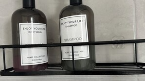 Twee flessen shampoo op een zwart schap tegen een betegelde muur.