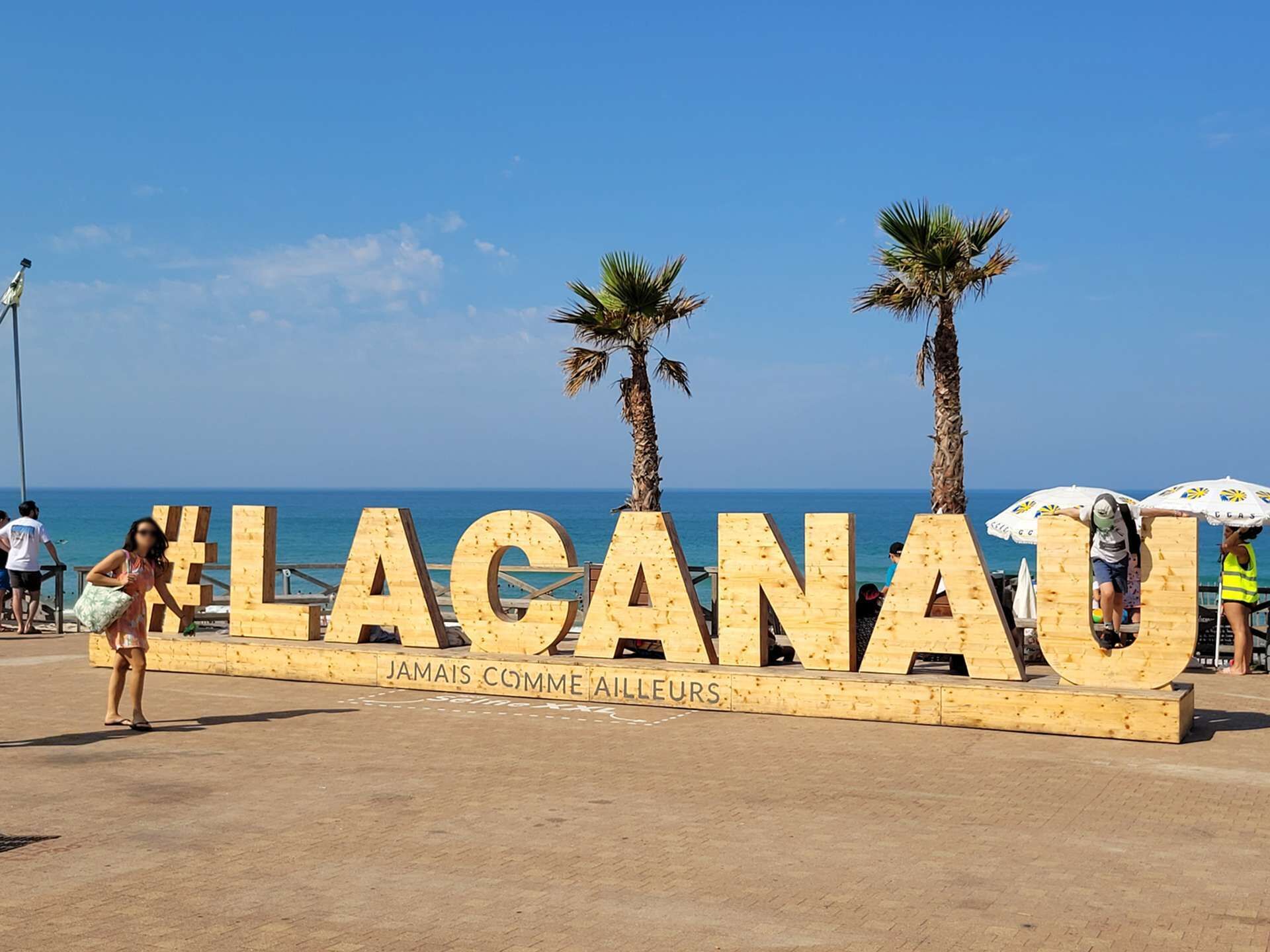 Ein hölzernes Schild mit dem Text 'LAGANAU' und 'JAMAIS COMME AILLEURS' am Strand, im Hintergrund Palmen und das Meer.