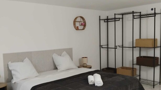 A modern hotel room with a bed, a bedside table, a wall-mounted shelf, and a hanging rack.