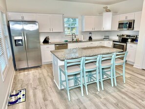 Eine Küche mit weißen Schränken, einem Edelstahl-Kühlschrank, einer Granit-Arbeitsplatte und drei blauen Stühlen.