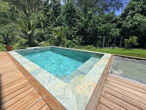 Ein rechteckiger Swimmingpool mit klarem, blauem Wasser, umgeben von einer Holzterrasse und üppiger Vegetation.