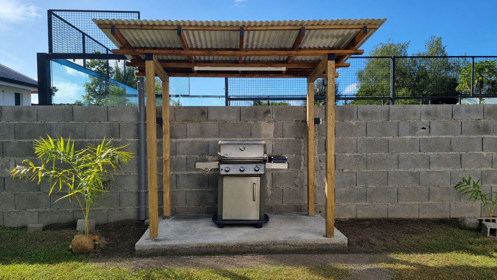 A barbecue grill under a wooden pergola with a brick wall backdrop.