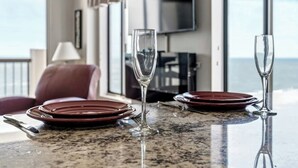 A dining table with plates and glasses, a television in the background, and a view of the ocean. - Oceanfront Corner Unit-Crescent Towers I 804-Pool (North Myrtle Beach)