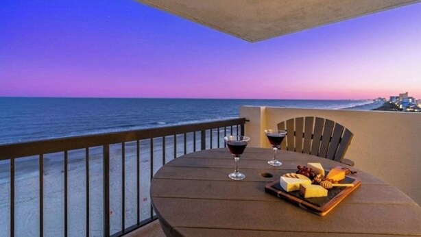 A balcony with a table set for a meal, overlooking the ocean at sunset. - Oceanfront Corner Unit-Crescent Towers I 804-Pool (North Myrtle Beach)
