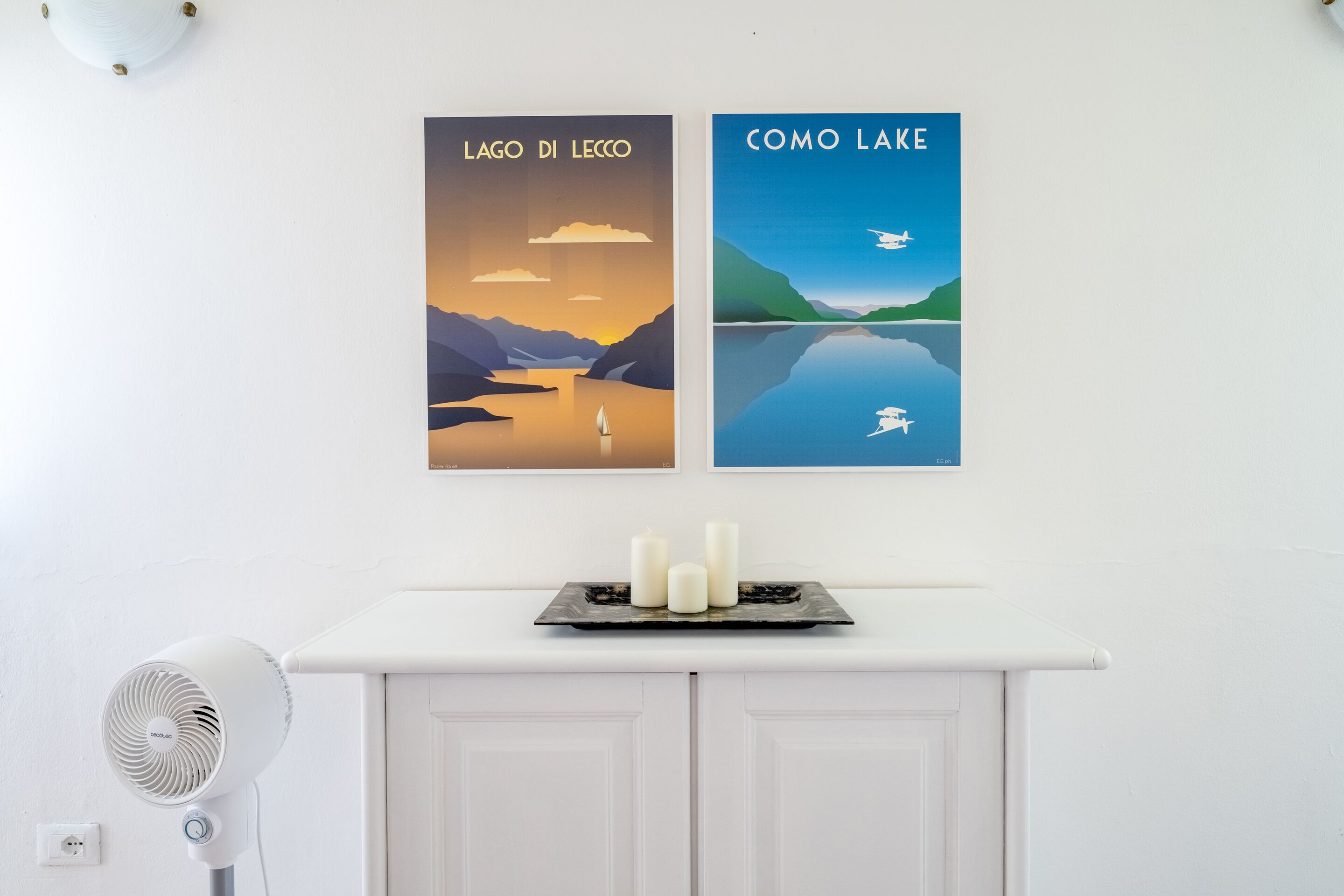Eine Wand mit zwei gerahmten Postern mit den Aufschriften 'LAGO DI LECCO' und 'COMO LAKE', eine weiße Kommode mit einer Ablage für drei Kerzen und ein weißer Ventilator links.