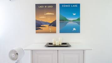 Eine Wand mit zwei gerahmten Postern mit den Aufschriften 'LAGO DI LECCO' und 'COMO LAKE', eine weiße Kommode mit einer Ablage für drei Kerzen und ein weißer Ventilator links.